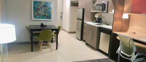 A modern kitchen with a refrigerator, microwave, and coffee maker. There is a dining area with a table and chairs, and a painting on the wall.