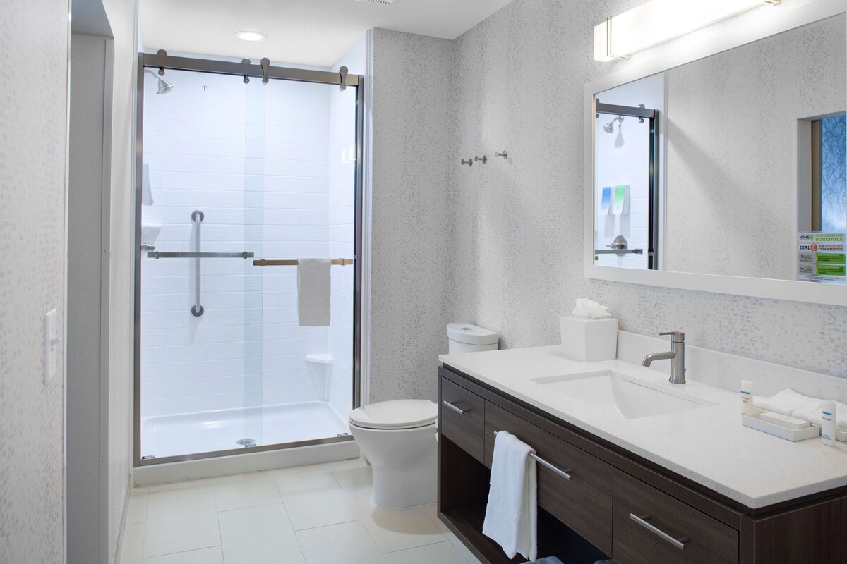 Un baño moderno con ducha envuelta en vidrio, inodoro, lavamanos con espejo y tocador con cajones.