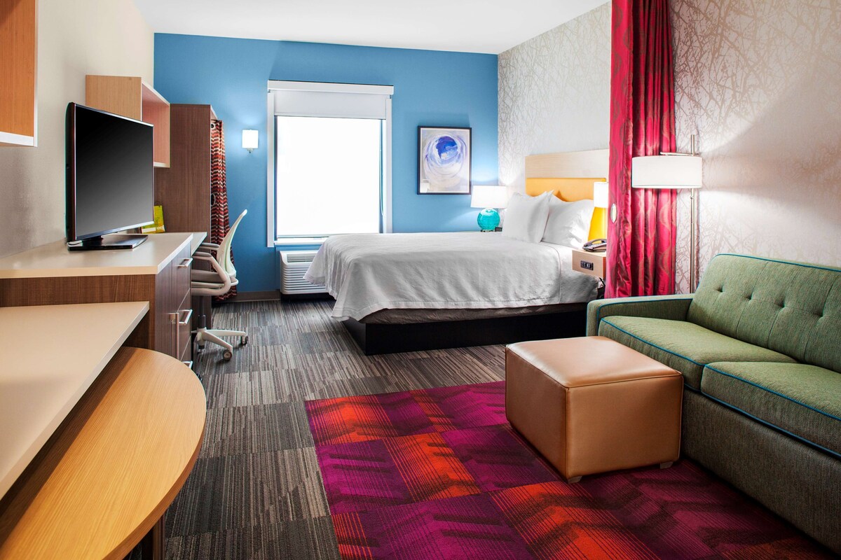 A hotel room with a bed, a desk, a TV, a sofa, and a window with curtains.