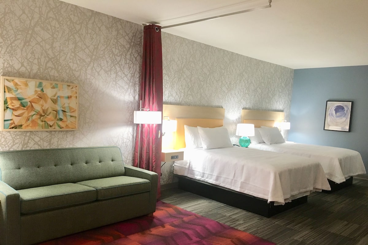 Een hotelkamer met een groene bank, een bed met witte lakens, een kamerbreed tapijt met dessin en een schilderij aan de muur.