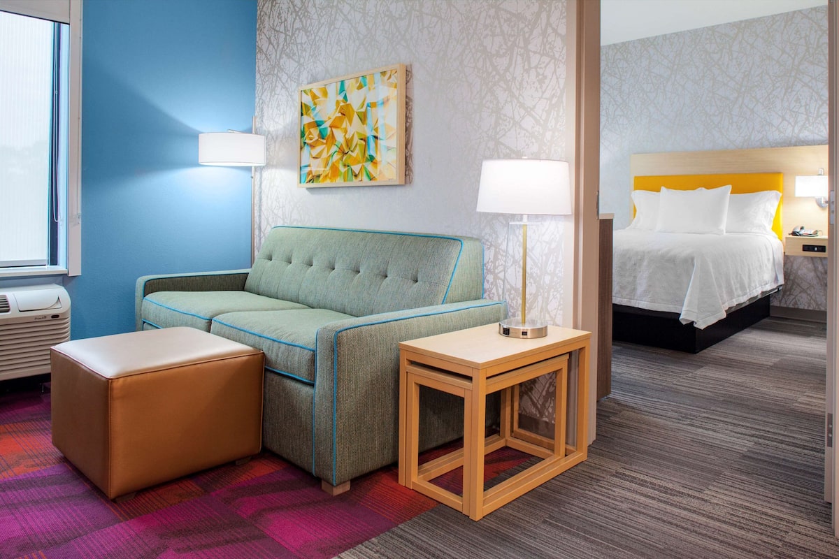 Een hotelkamer met een groene bank, een houten nachtkastje, een bed met witte beddengoed en een kleurrijk vloerkleed.