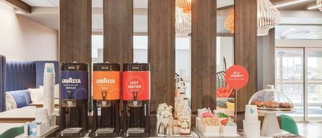 A hotel reception area with a coffee station featuring Lavazza and Hot Water coffee machines, a glass display case with snacks, and a sign indicating a hurry service.