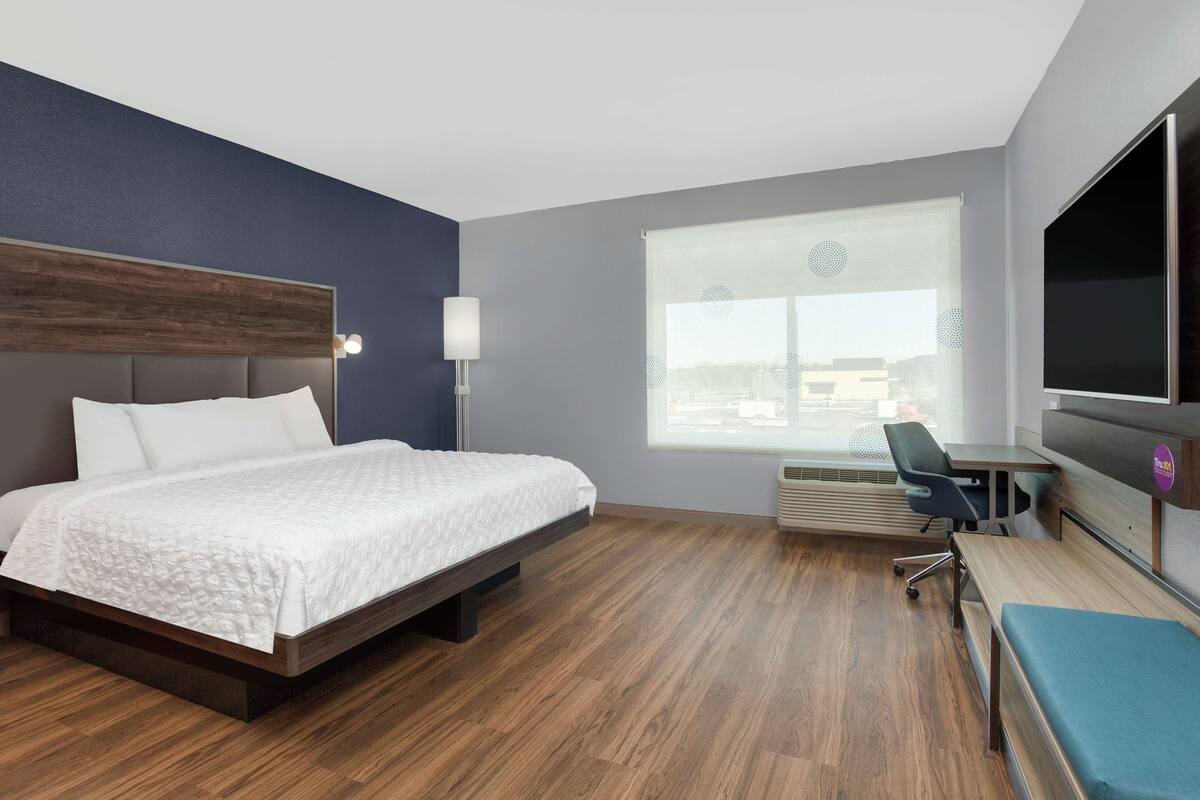 Une chambre d’hôtel moderne avec un lit en bois, un bureau et une grande fenêtre équipée de stores.