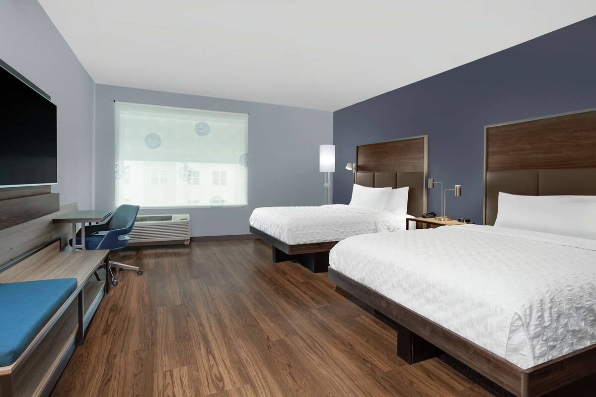 Une chambre d’hôtel avec deux lits, un bureau, une chaise, une télévision et une fenêtre avec des stores.