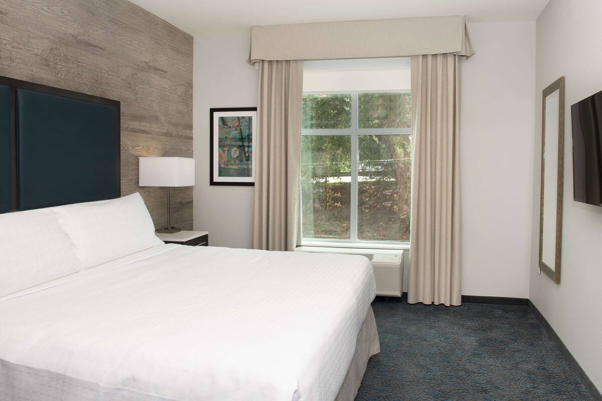 Ein Hotelzimmer mit einem großen Bett, einem Fenster mit Vorhängen, einem gerahmten Bild an der Wand und einem Flachbildfernseher.