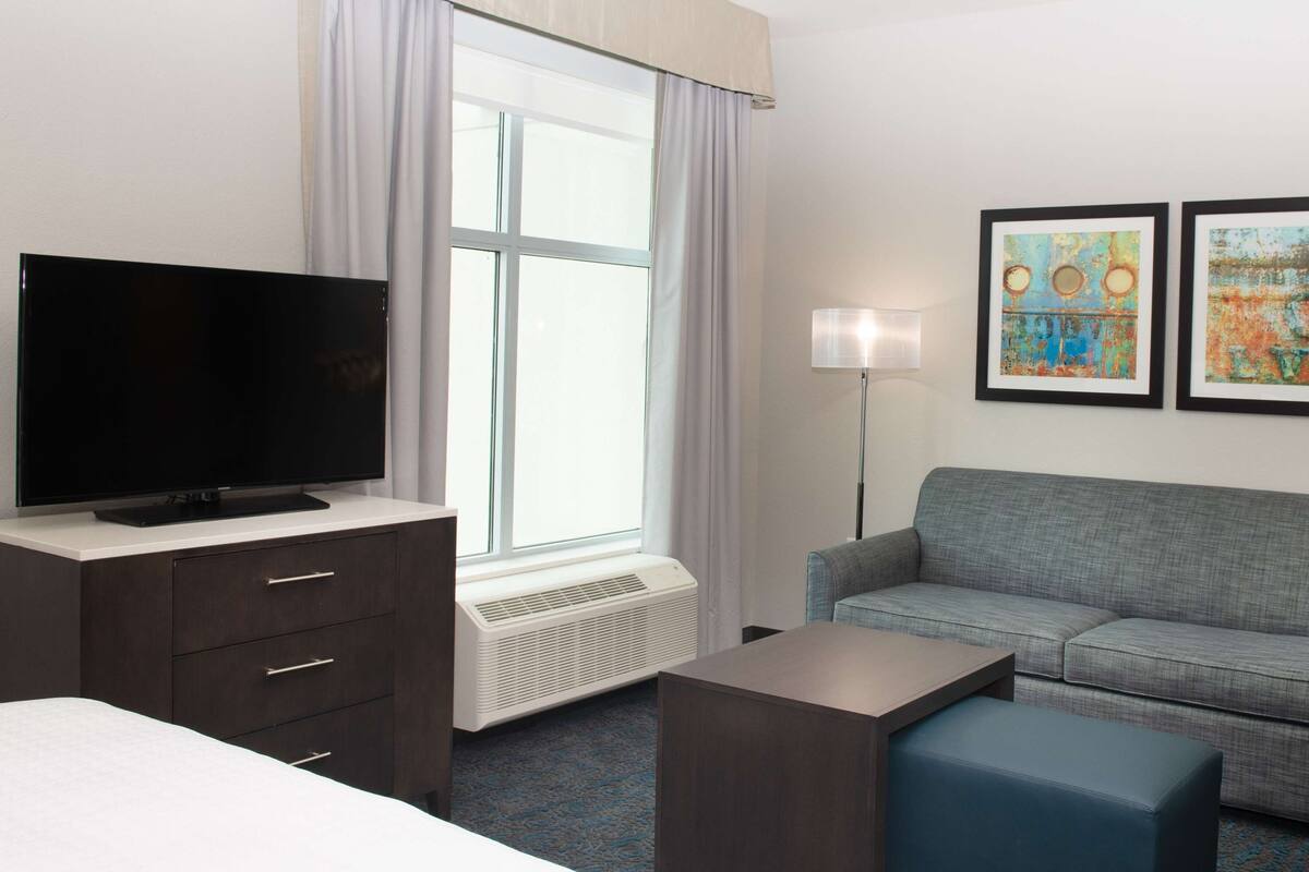 Een hotelkamer met een flatscreen-tv, een grijze bank, een blauwe fauteuil, een klein tafeltje en twee kunstwerken in lijsten aan de muur.