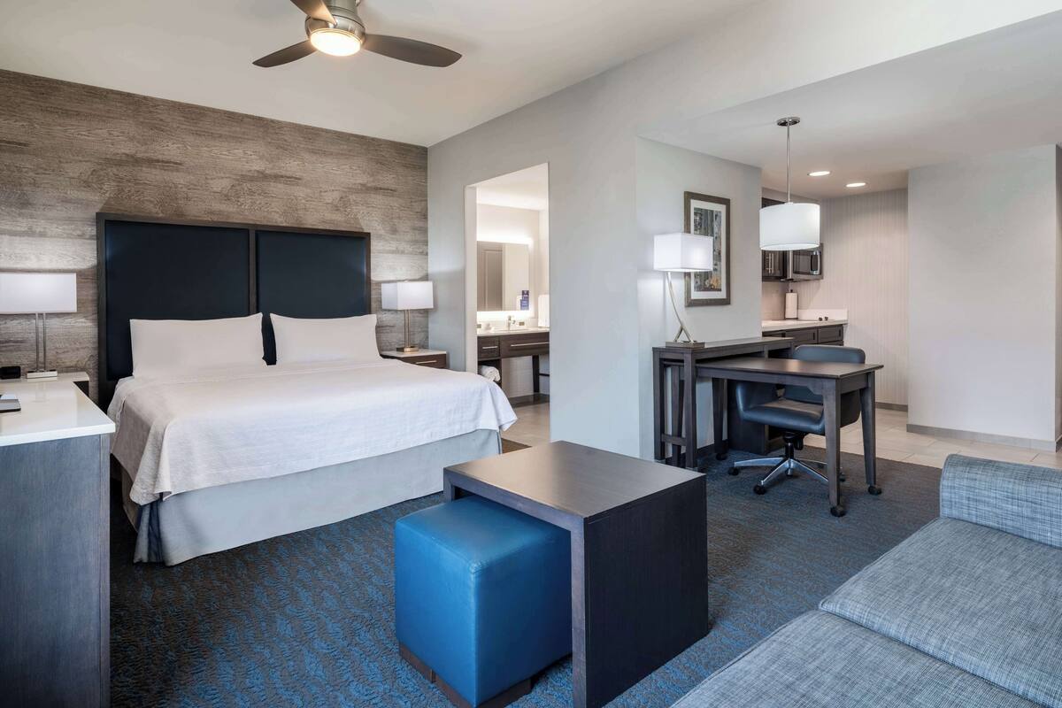Een hotelkamer met een bed, bureau, stoel en een klein blauw voetenbankje.