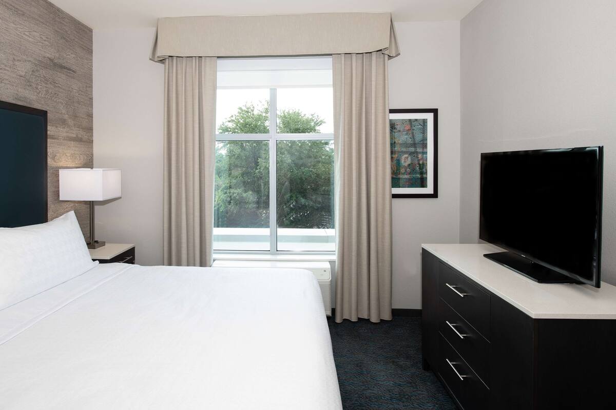 Ein Hotelzimmer mit Bett, Fernseher auf einer Kommode, einem Fenster mit Vorhängen und einem gerahmten Bild an der Wand.