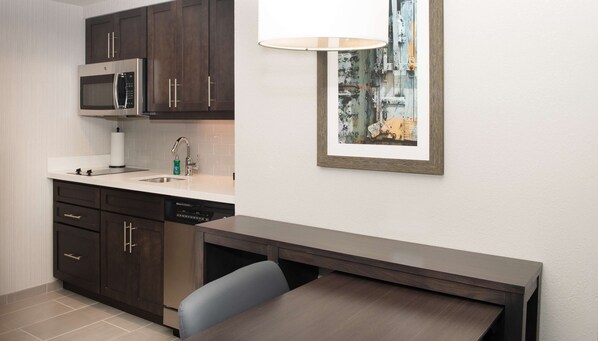 Una cocina compacta con armarios oscuros, un microondas, un lavamanos y una zona de comedor con mesa y silla.