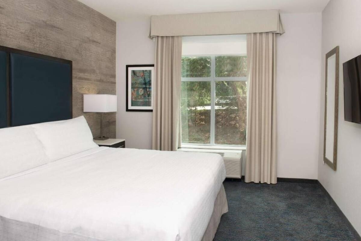 Een hotelkamer met een bed, een raam met gordijnen, een aan de muur bevestigde televisie en een lijst met een afbeelding.