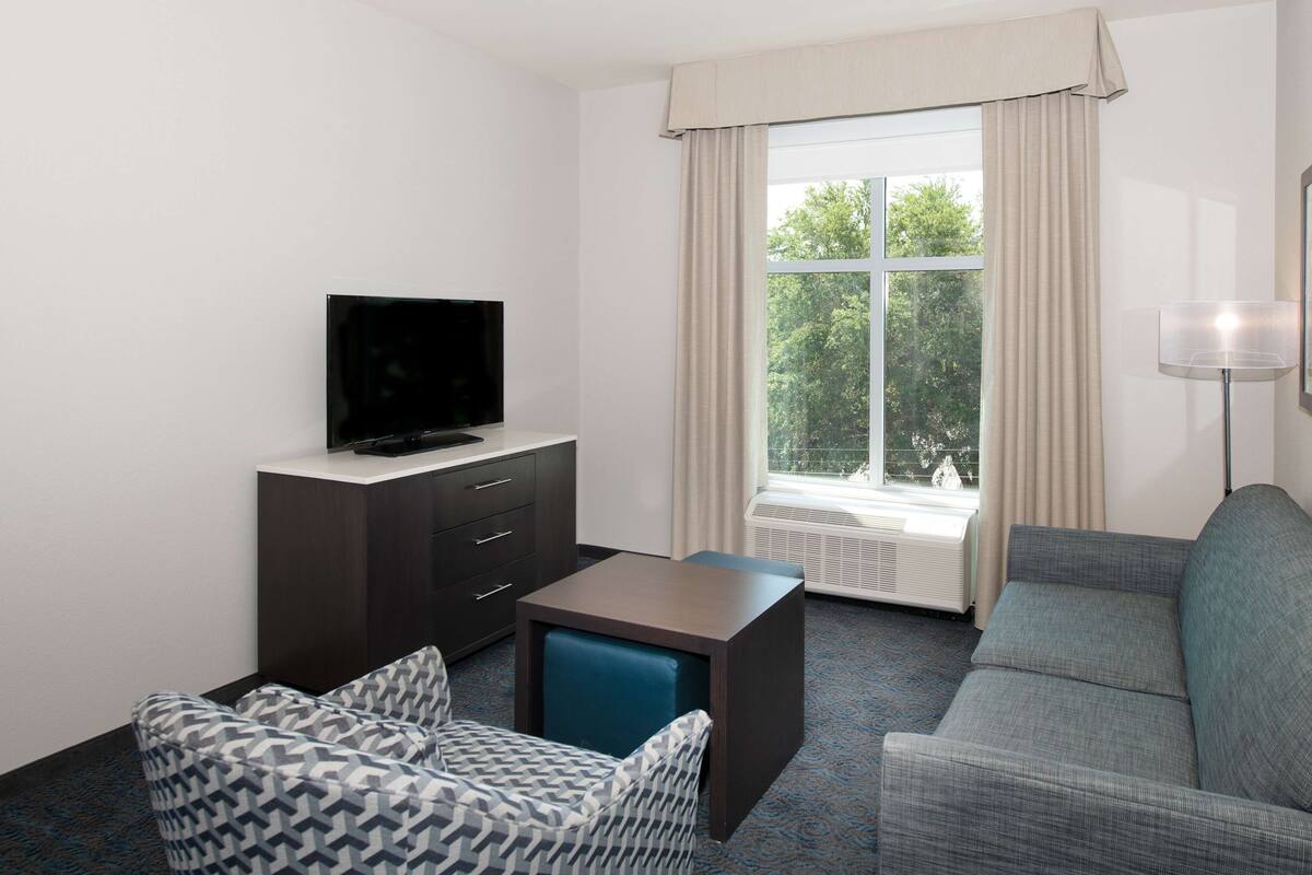 Ein modernes Hotelzimmer mit Fernseher auf einer dunklen Holzkommode, einem grauen Sofa, einem gemusterten Sessel und einem großen Fenster mit Vorhängen.
