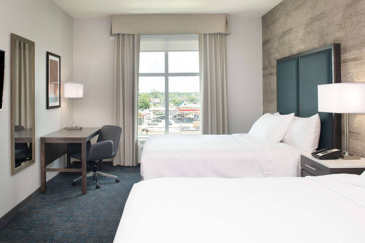 Ein Hotelzimmer mit zwei Betten, einem Schreibtisch, einem Stuhl, einem Fernseher und einem Fenster mit Blick auf Gebäude.