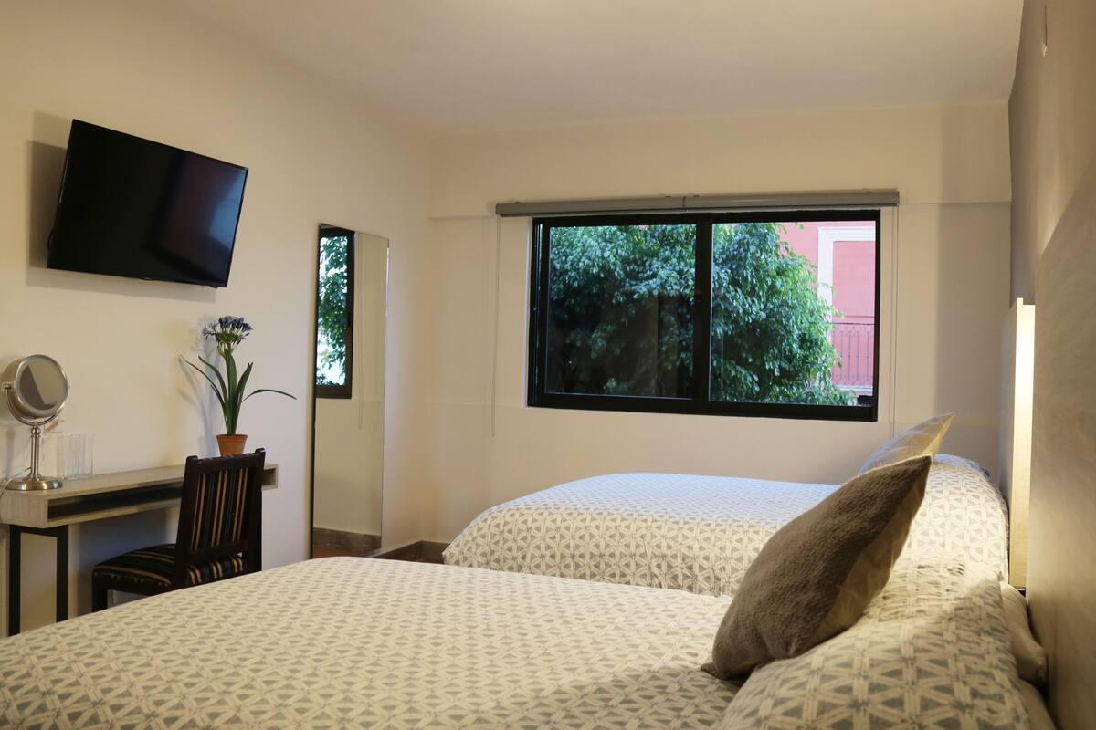 A hotel room with a bed, a desk with a chair, a TV mounted on the wall, and a window with a view of trees and a building.