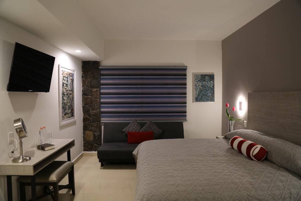 A modern bedroom with a bed, a desk, a bench, a TV, and artwork on the walls.
