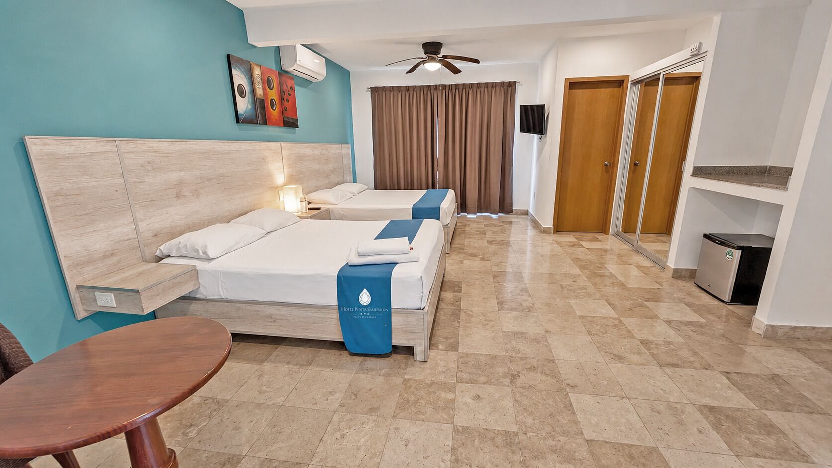 A hotel room with two beds, a desk, a chair, a ceiling fan, and a TV.
