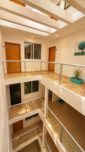 Interior de hotel con varios niveles, barandillas de vidrio, puertas de madera y una planta en maceta.