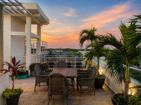 Una terraza en la azotea con mesa y sillas, plantas en macetas y vistas panorámicas del horizonte urbano al atardecer.