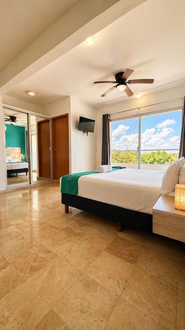 Habitación de hotel con cama, ventilador de techo, televisión y un gran ventanal con vistas.