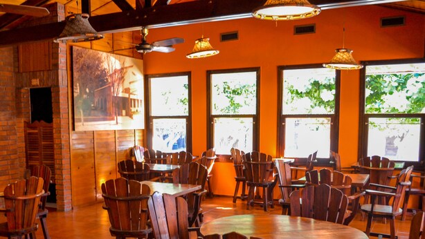 A restaurant with wooden tables and chairs, large windows with greenery outside, and hanging lamps. - Hotel Cheval Blanc (Villa Gesell)