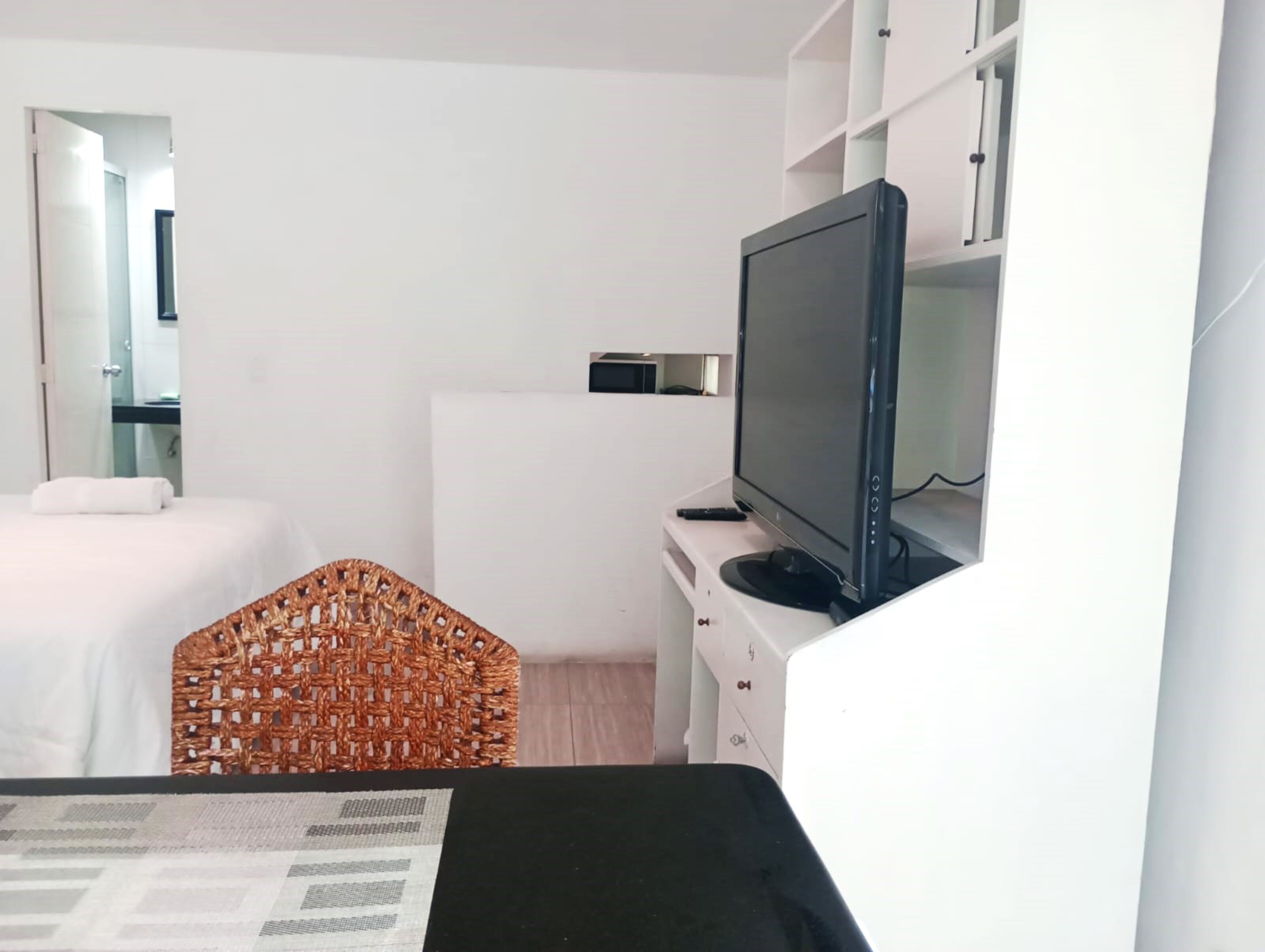 Habitación de hotel con cama, televisor sobre un mueble bajo, una mesita pequeña y una silla de mimbre.
