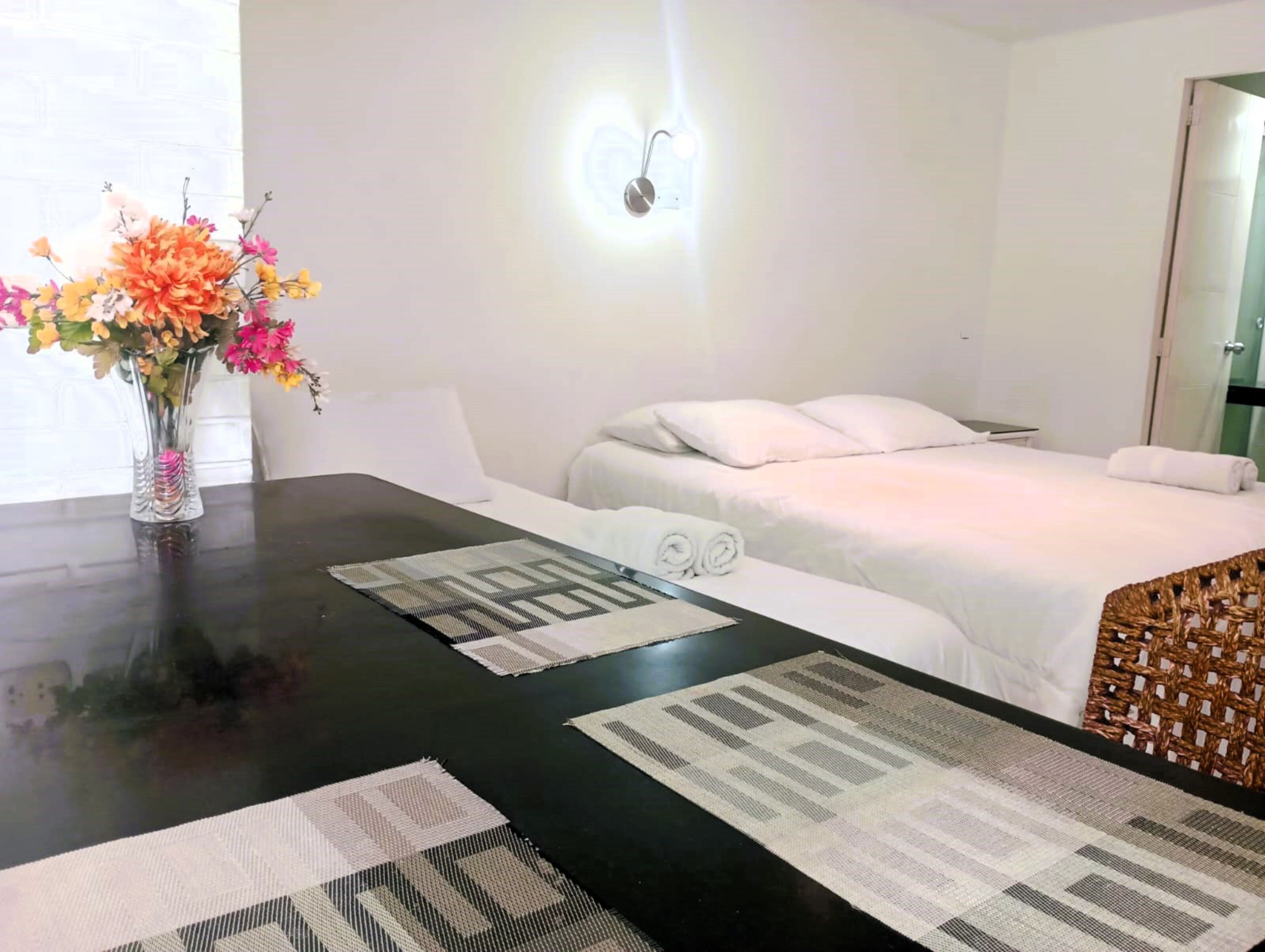 A hotel room with a bed, a bedside table with a vase of flowers, and a wall-mounted light fixture.