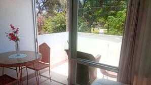 A room with a bed, a round table with a vase of flowers, a wicker chair, and a view of a park through a large window.