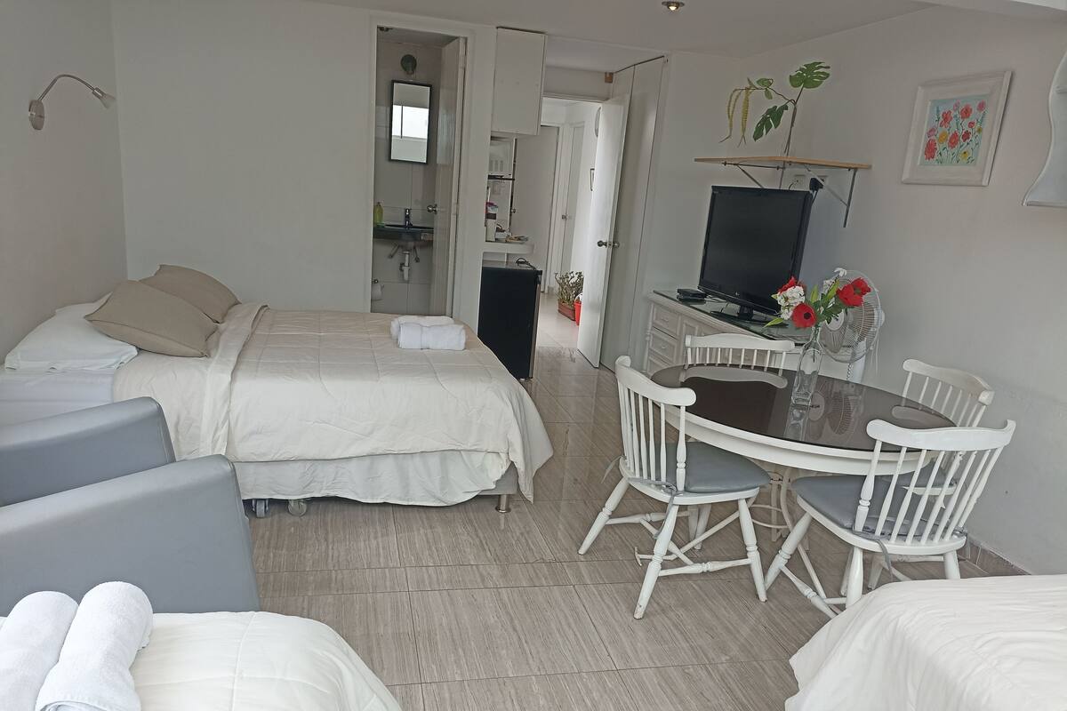 A hotel room with two beds, a dining table with chairs, a TV, and a small kitchen area.