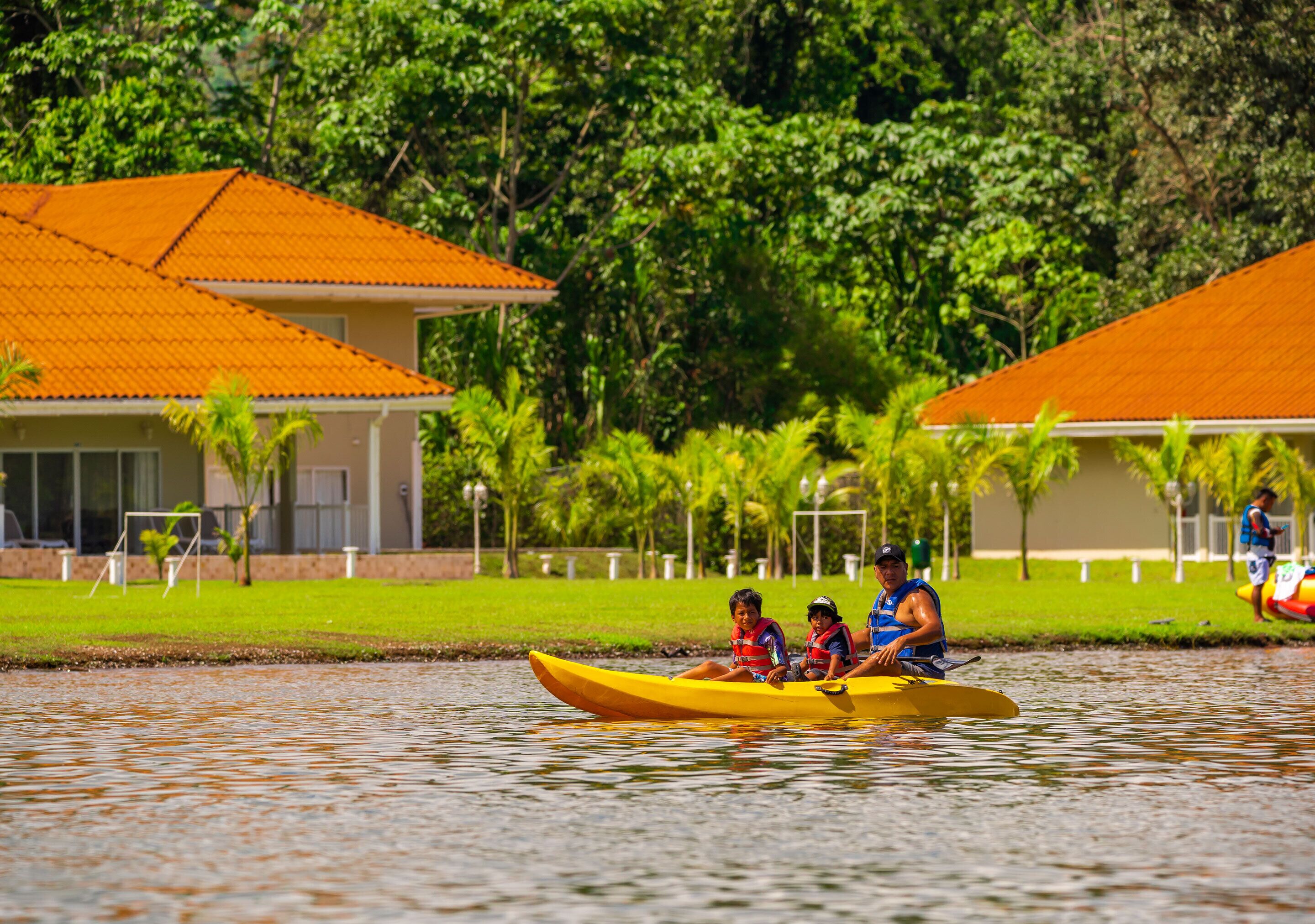 A yellow kayak with three people on a calm body of water, surrounded by lush greenery and buildings with orange roofs.