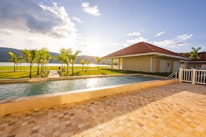 A swimming pool area with a tiled deck, a covered structure, and a view of a body of water and mountains in the background.
