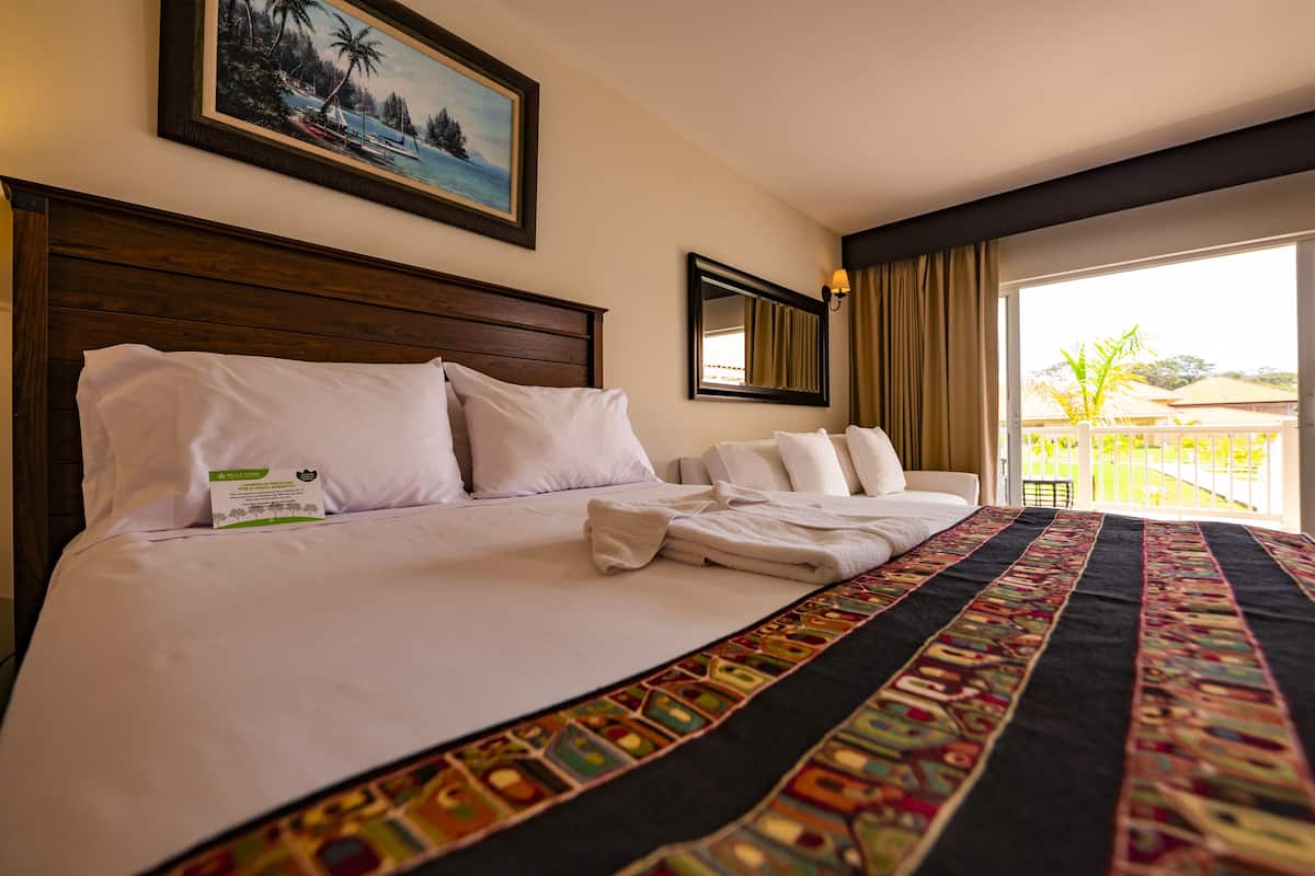 A hotel room with a large bed, a painting of a beach scene, a window with curtains, and a balcony with a view of palm trees.