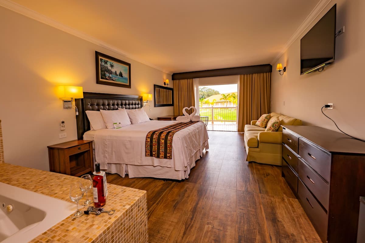 A hotel room with a bed, a desk with a lamp, a TV, a sofa, and a balcony with a view.