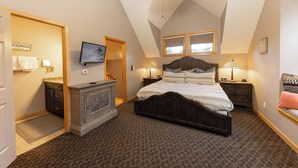 Een slaapkamer met een schuine kap, een bed met houten hoofdbord, twee nachtkastjes met lampen, een televisie aan de muur en een raam met uitzicht.