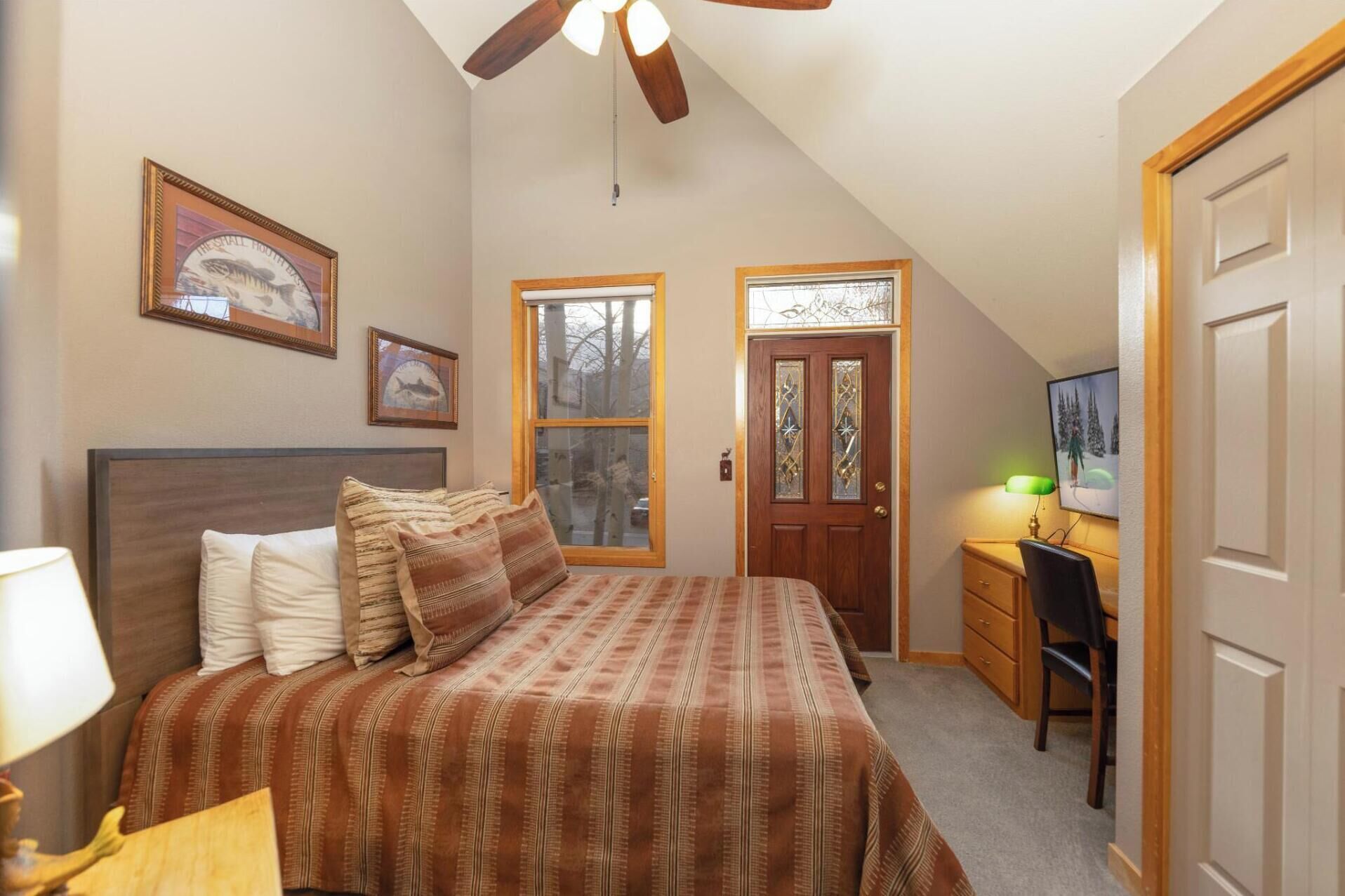 A bedroom with a bed, a desk, a chair, a ceiling fan, and two windows.