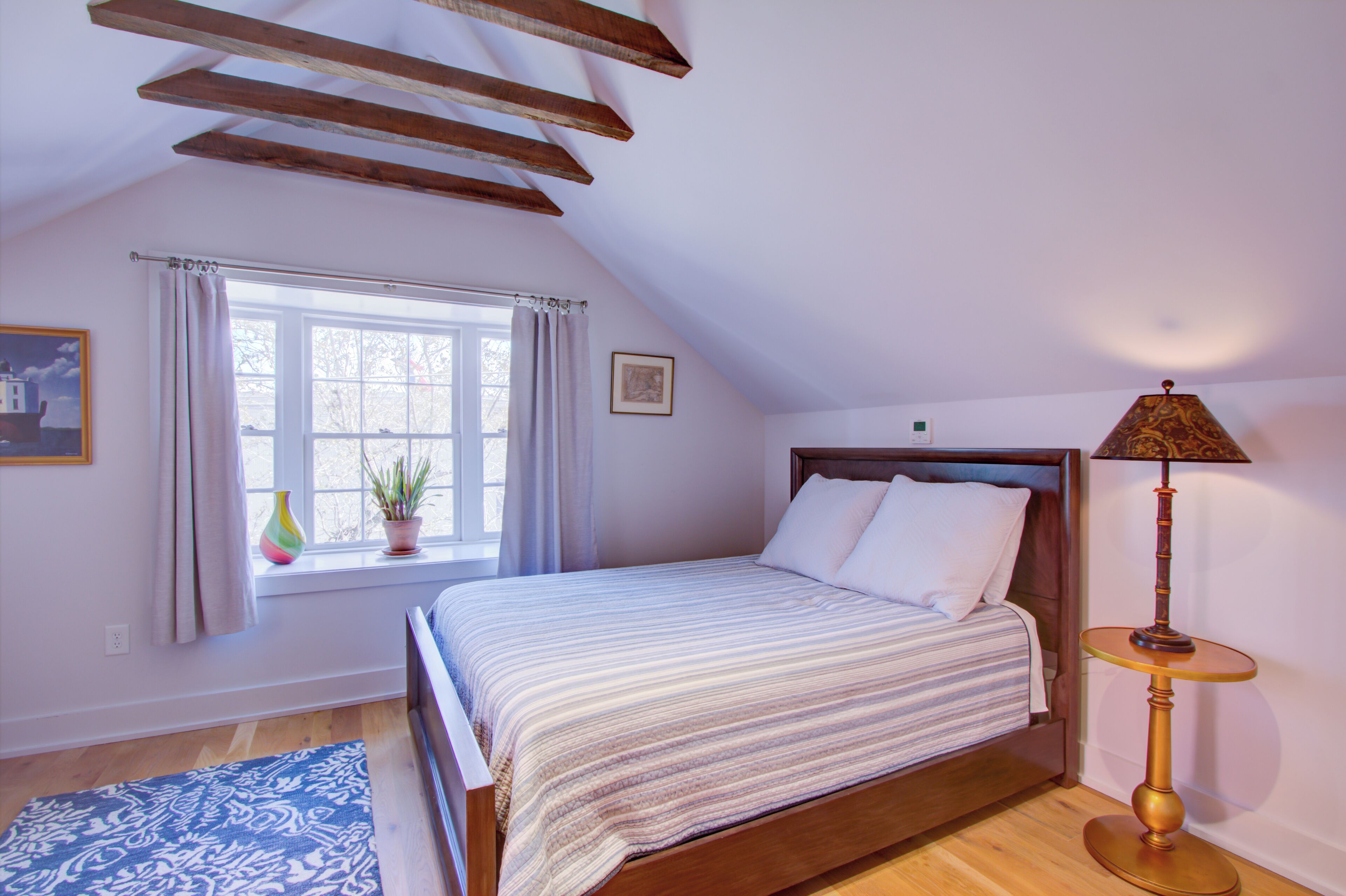 A bedroom with a bed, a window with curtains, a bedside table with a lamp, and a small potted plant.