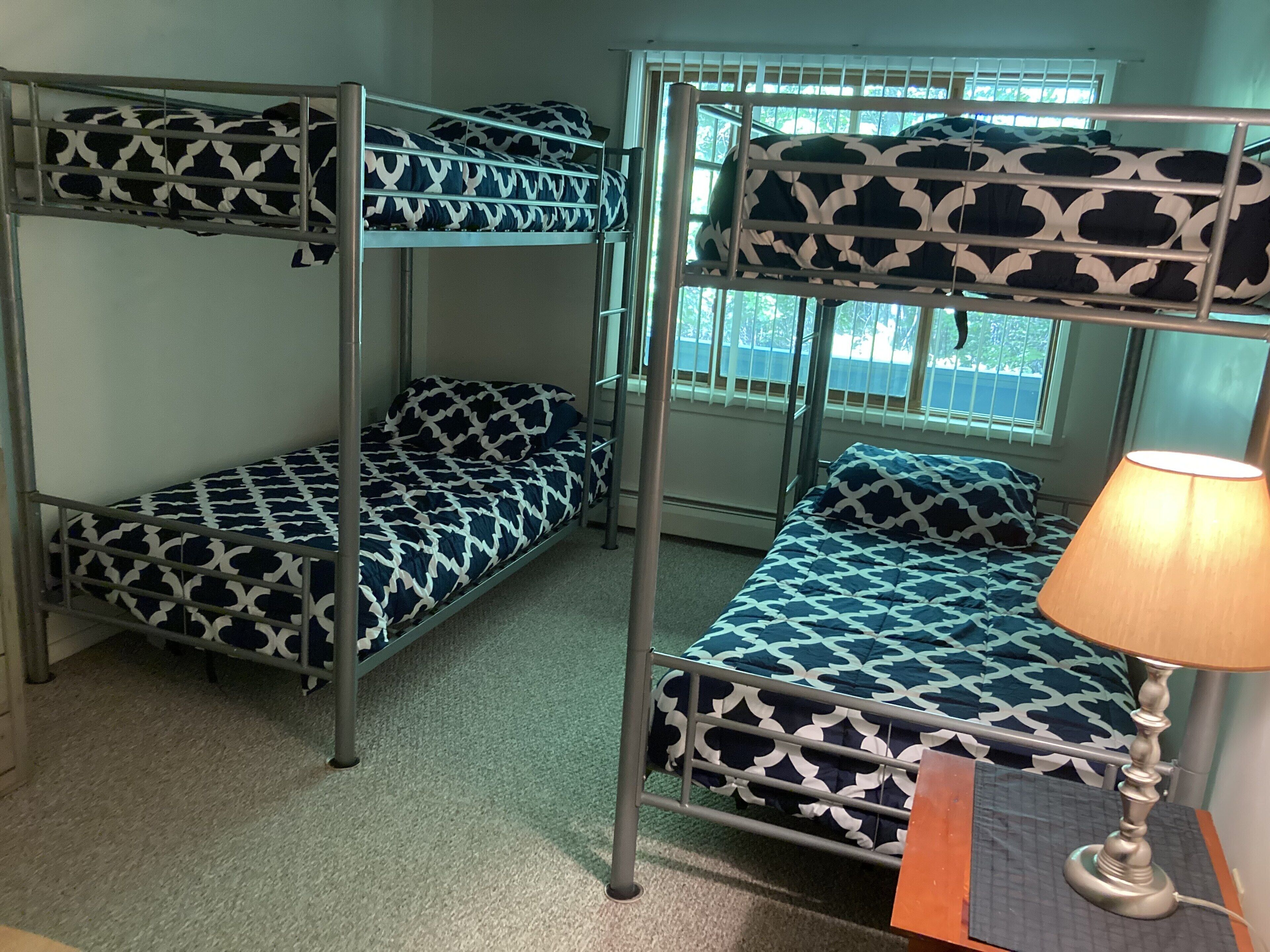 A room with two bunk beds, each with a patterned bedspread, a bedside table with a lamp, and a window with blinds.