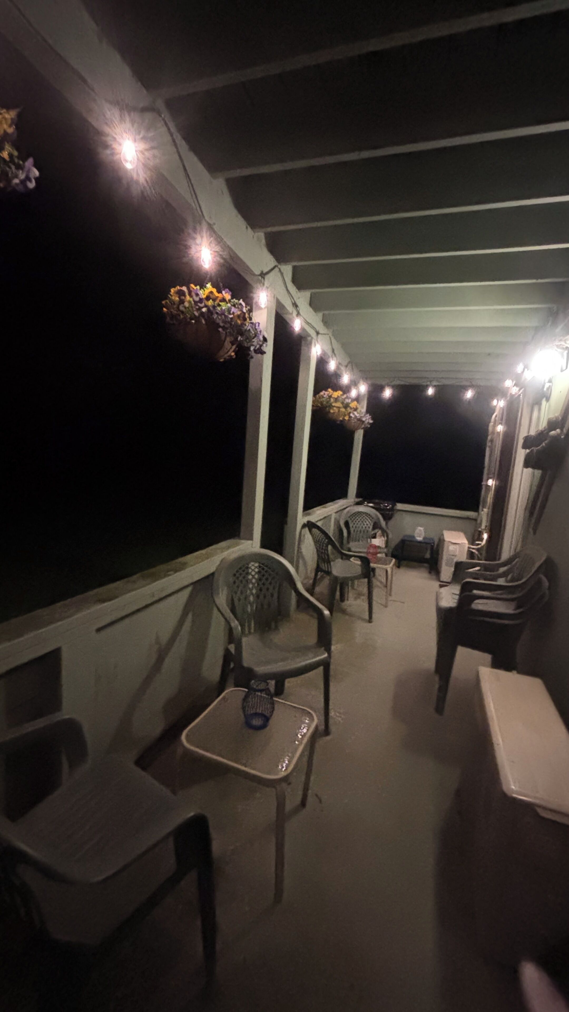 Un área de estar al aire libre con techo, sillas y mesas, luces colgantes y cestas de flores.
