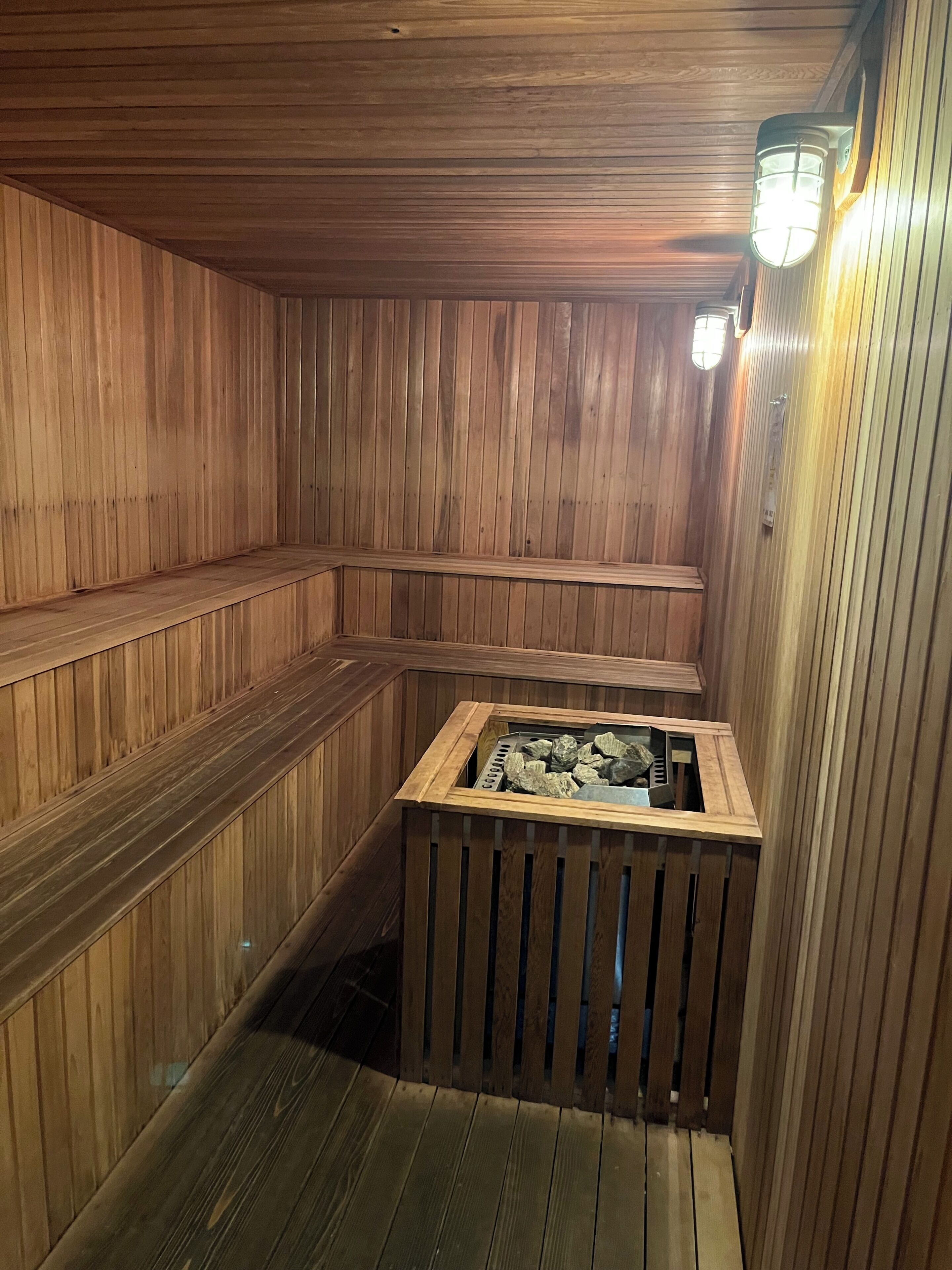 A sauna with wooden walls and benches, a central heated stone feature, and a wall-mounted light fixture.