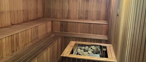 A sauna with wooden walls and benches, a central heated stone feature, and a wall-mounted light fixture.