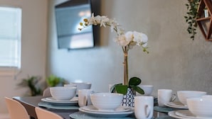 Une table à manger dressée avec de l’assiette, des bols et des tasses blancs, et une orchidée blanche dans un vase.