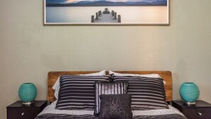 Une chambre à coucher avec une tête de lit en bois, une literie à rayures et deux vases turquoise placés sur les tables de chevet. Un tableau encadré représentant une jetée s’avançant dans un lac est accroché au mur.