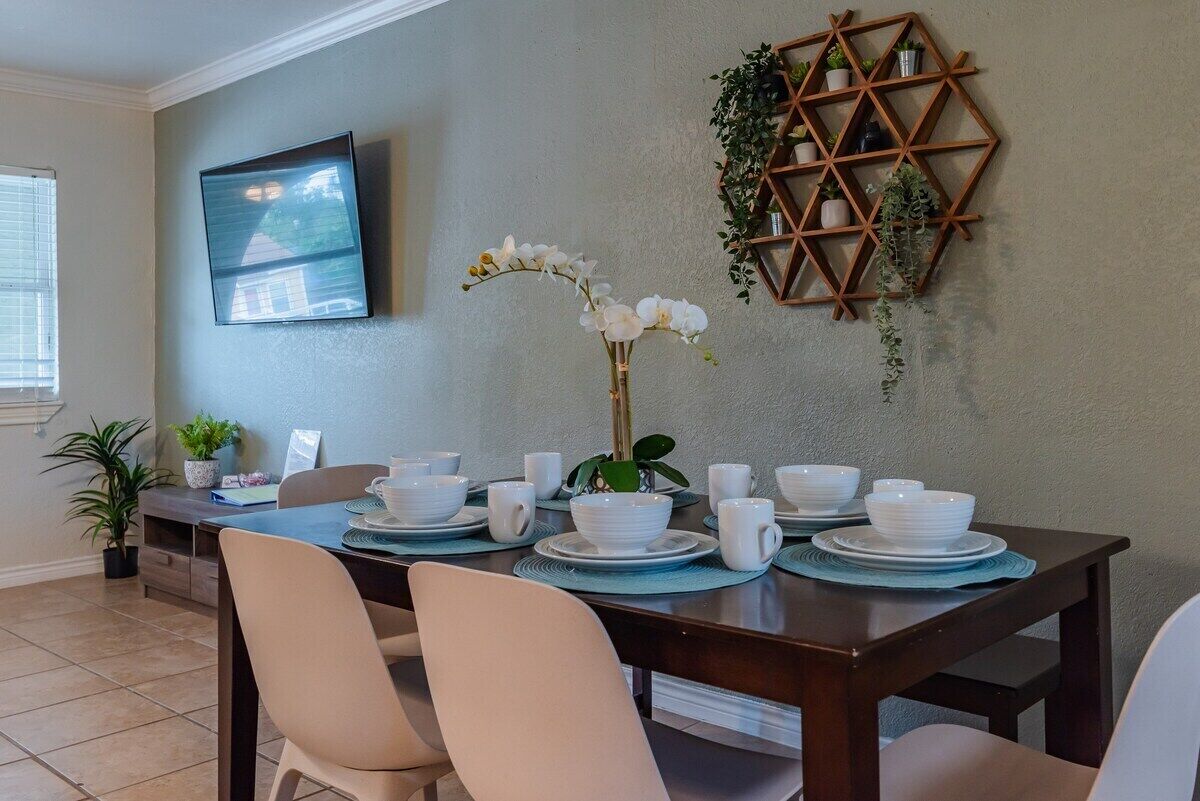A dining area with a dark wooden table set for four, surrounded by beige chairs. A wall-mounted TV is visible on the left, and a geometric wooden shelf with plants is on the right.