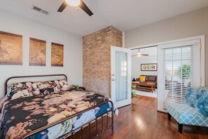Un dormitorio con cama, pared de ladrillo, puerta que da a una sala de estar y ventana con persianas.