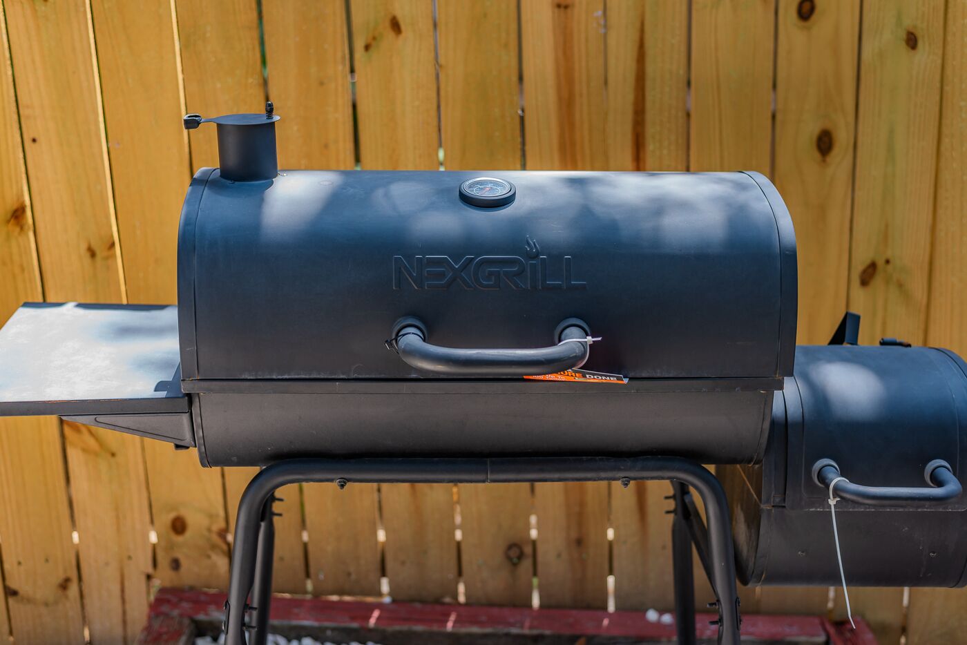 A black NEXGRILL barbecue grill with a handle and a lid, set against a wooden fence background.