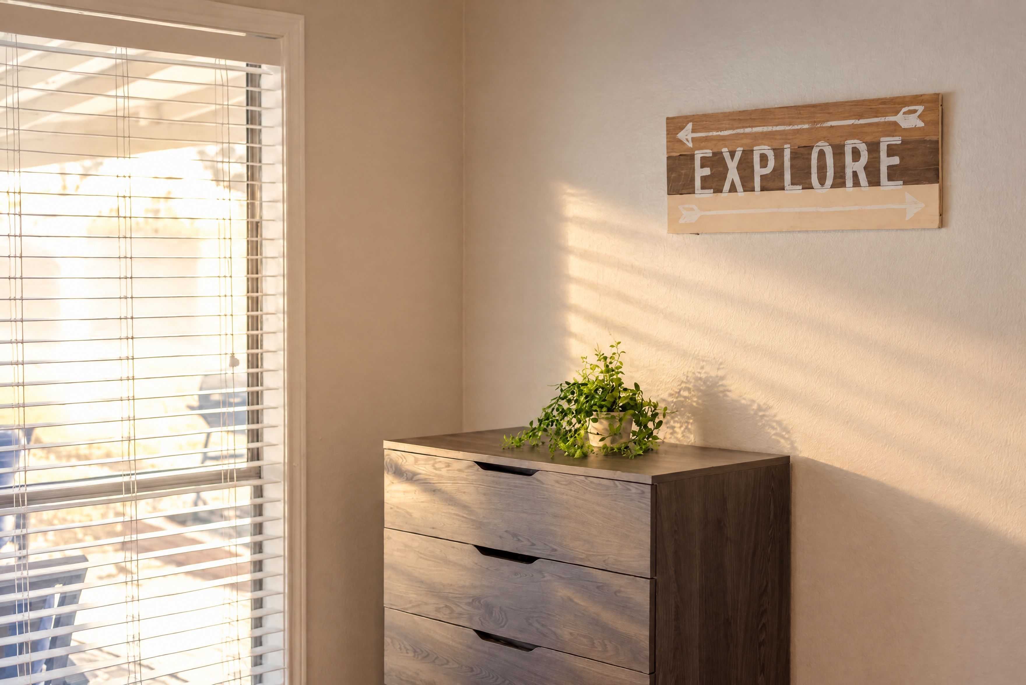 Quarto com janela e persianas, armário de madeira com gavetas e um aviso na parede que diz 'EXPLORE'.