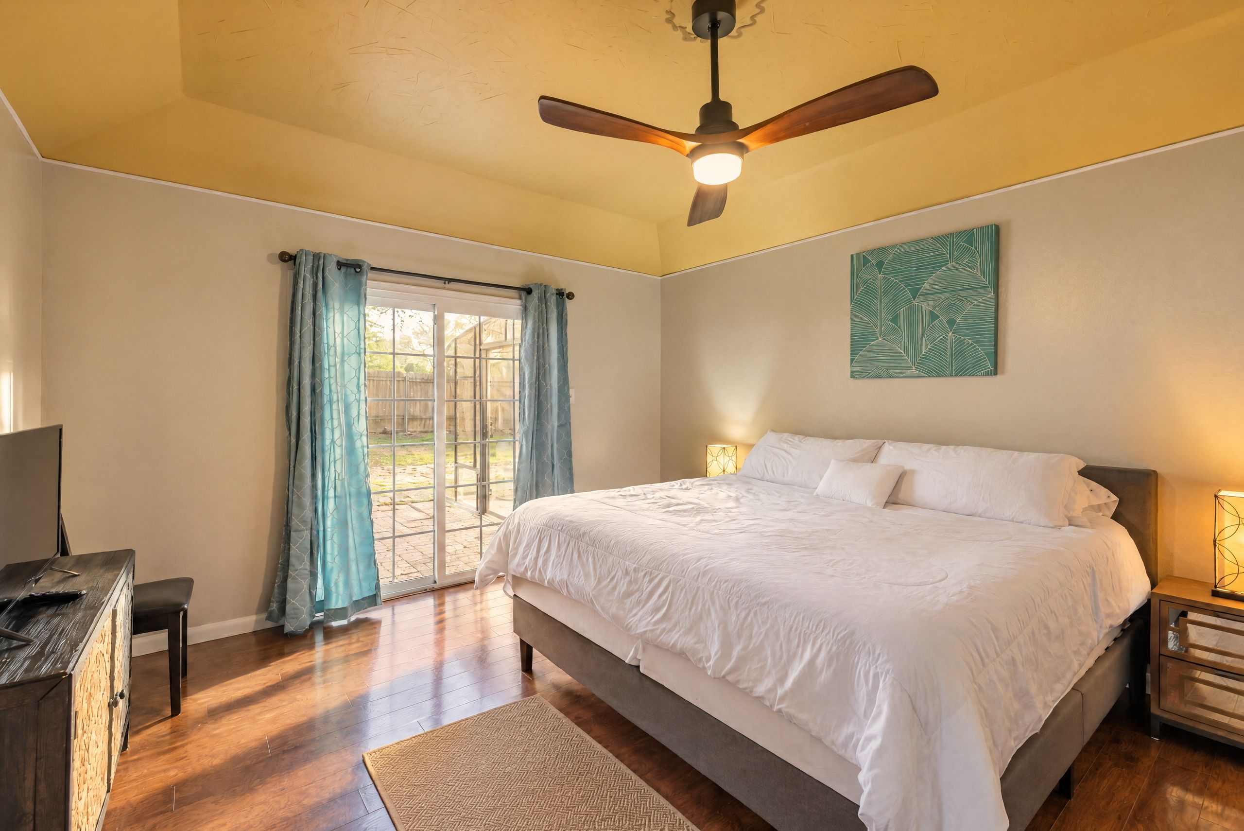 Une chambre à coucher comprenant un lit, un ventilateur de plafond, une fenêtre avec des rideaux et une œuvre d’art accrochée au mur.