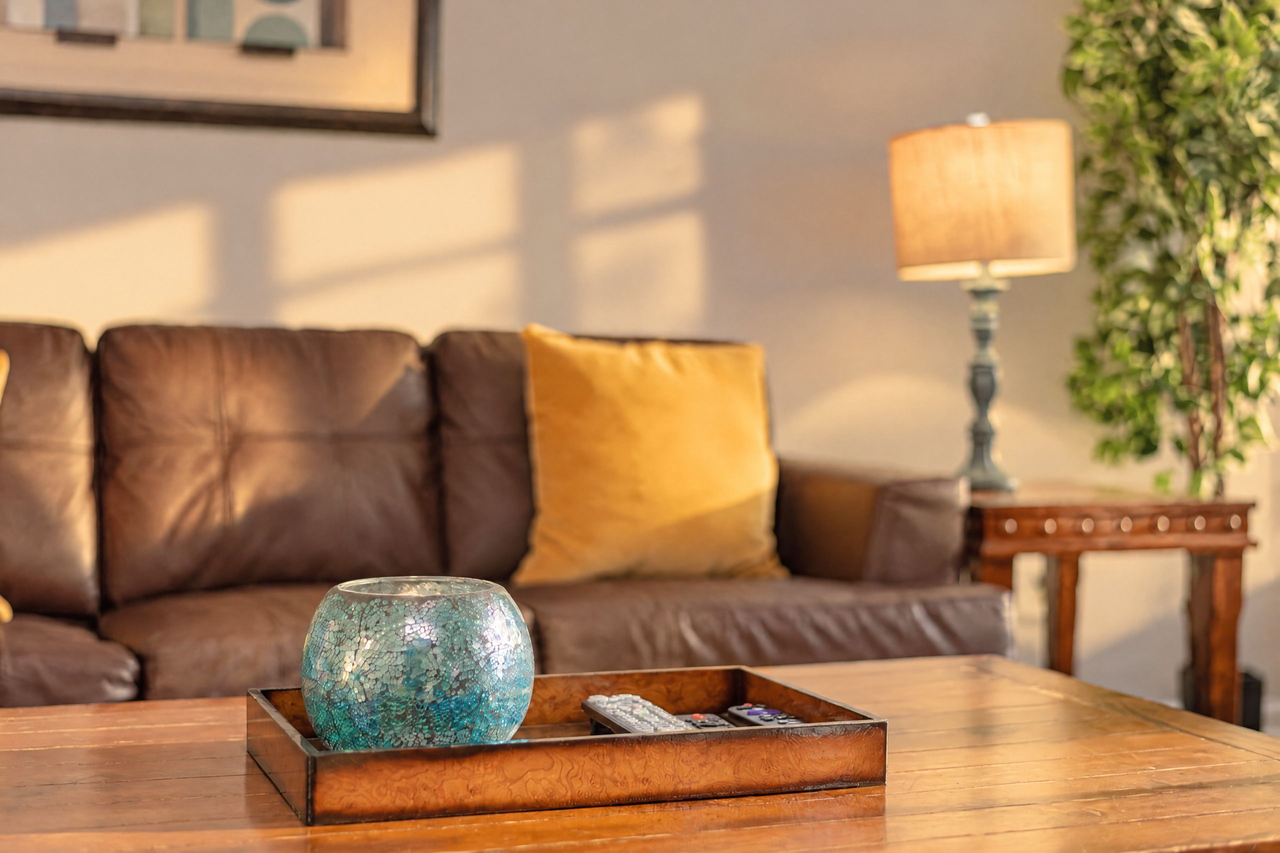 Ein Holztisch mit einer dekorativen Glasfigur und Fernbedienungen, ein braunes Ledersofa mit einem gelben Kissen, eine Lampe und ein gerahmtes Bild an der Wand.
