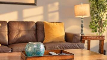 Ein Holztisch mit einer dekorativen Glasfigur und Fernbedienungen, ein braunes Ledersofa mit einem gelben Kissen, eine Lampe und ein gerahmtes Bild an der Wand.