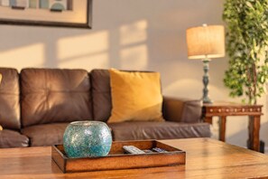 A wooden coffee table with a decorative glass object and remote controls, a brown leather sofa with a yellow cushion, a lamp, and a framed picture on the wall.