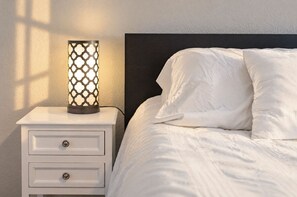 Una cama con ropa de cama y almohadas blancas, una mesita de noche con una lámpara y un cajón con pomo redondo.