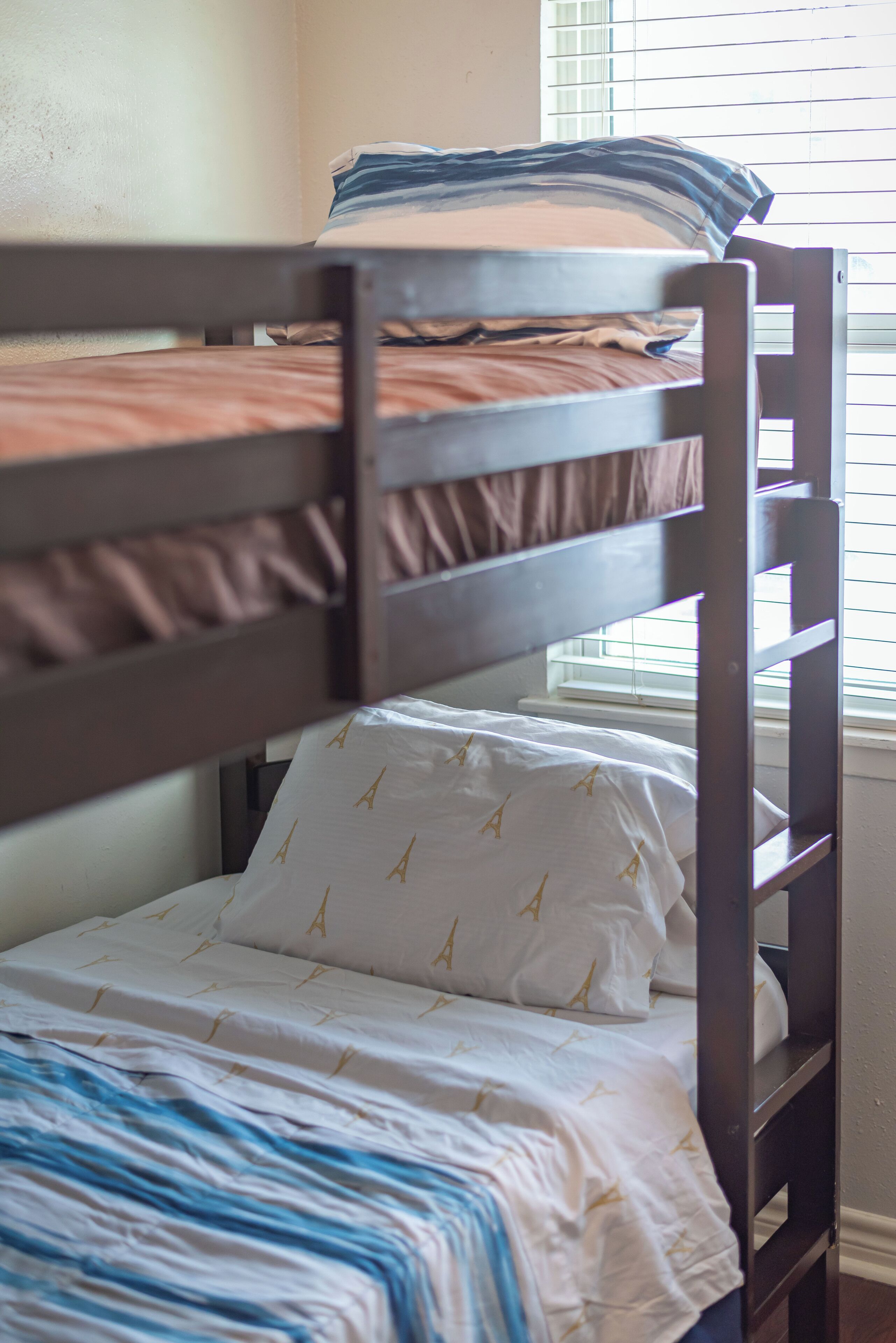 Una cama litera con colchón blanco y almohadas estampadas, cabecera de madera y una ventana con persianas.